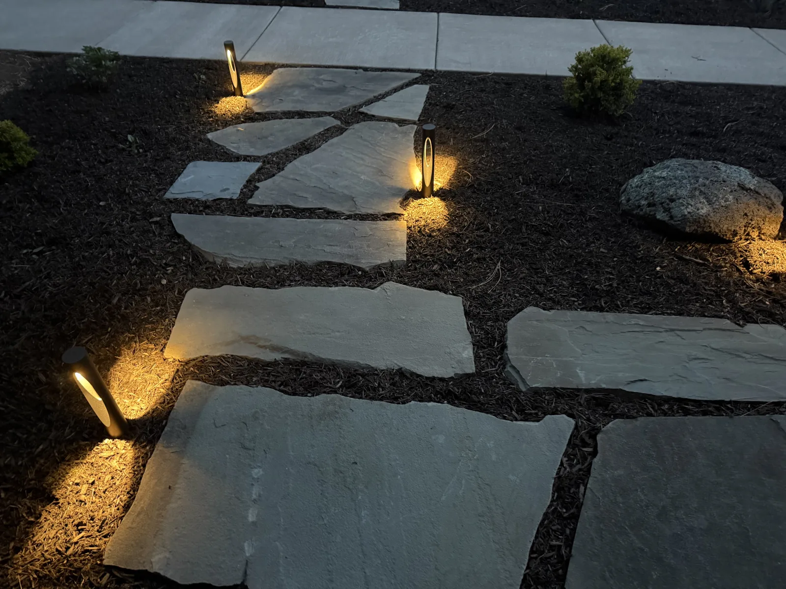 Close-up of low-voltage path lights illuminating a flagstone stepping stone walkway through dark mulch beds