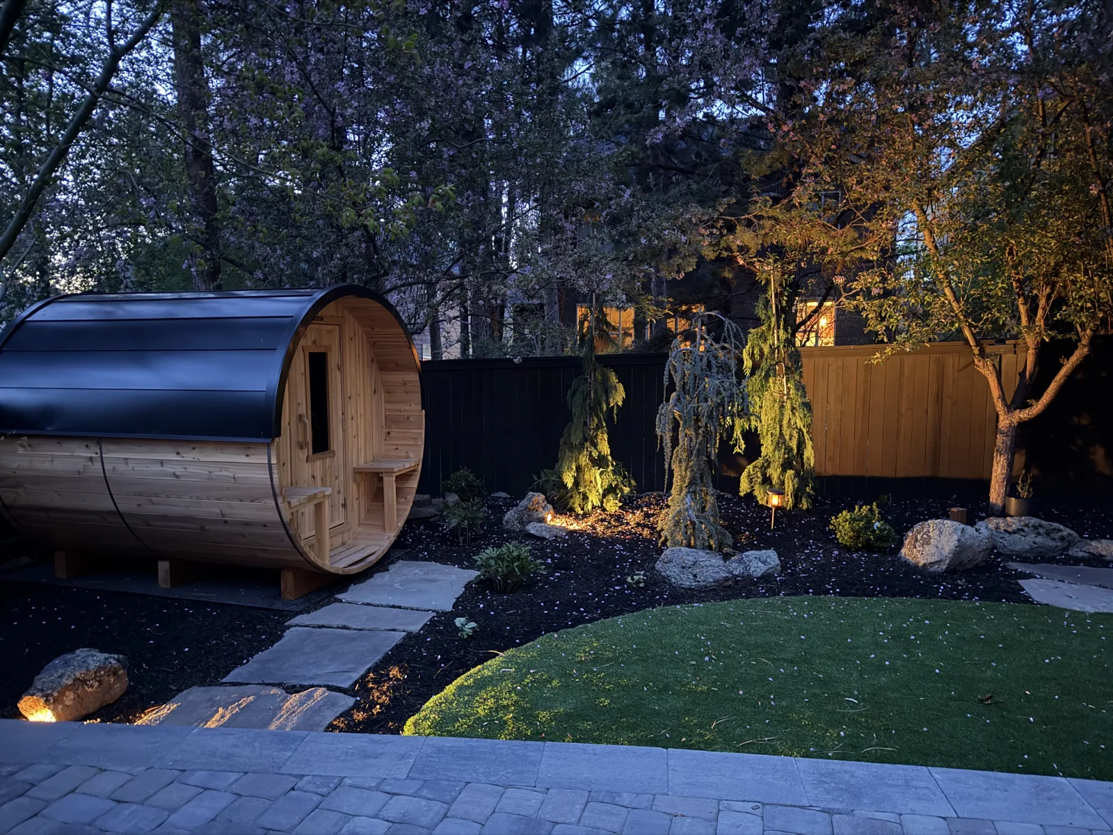 Backyard scene at dusk with uplighting on a weeping ornamental tree, cedar barrel sauna, and accent path lights