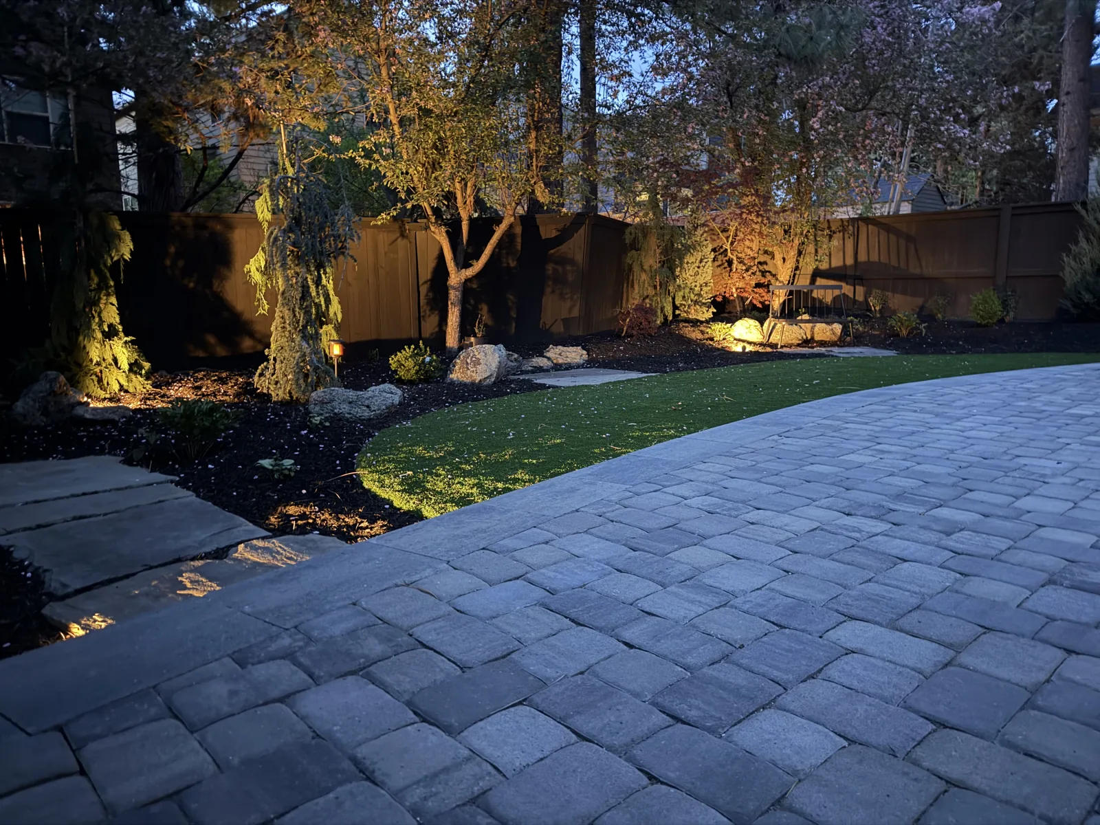 Curved paver patio at dusk with tree uplighting and warm accent lights along the planting beds