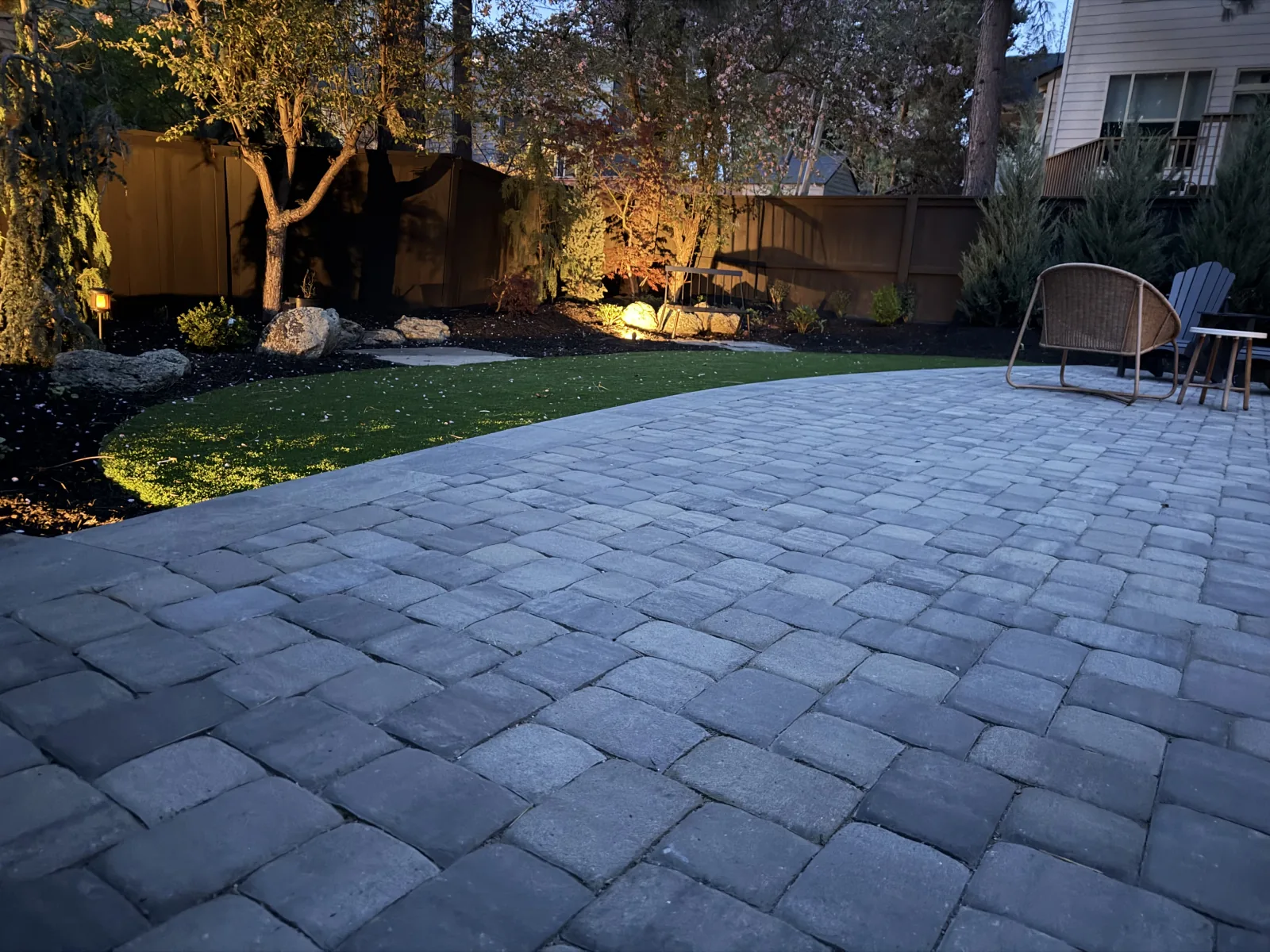 Large paver patio with outdoor seating, uplit trees, and warm landscape lighting at dusk